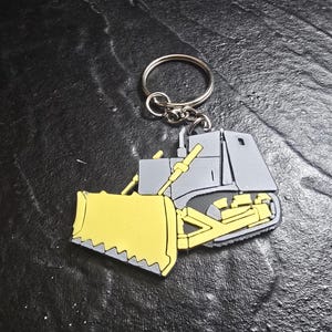 May include: A yellow and gray bulldozer keychain with a silver key ring. The bulldozer has a large yellow blade and a gray cab.