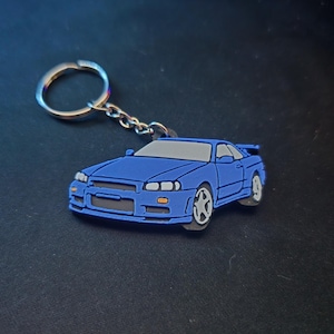 May include: A blue car keyring with a silver key ring and chain. The car has a grey roof and white wheels. The keyring is made of a rubbery material.