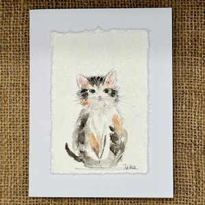 May include: Watercolor painting of a seated kitten with black, brown, and white fur. The artwork is on textured paper with a deckled edge, set within a white mat. The background is a burlap texture.