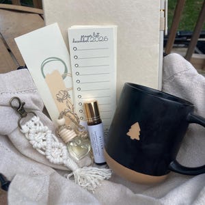 May include: A gift set featuring a black mug with a tree design, a journal, a prayer list dated December 2026, a macrame keychain, a roller bottle, and a small heart-shaped bottle. The items are arranged in a beige drawstring bag.