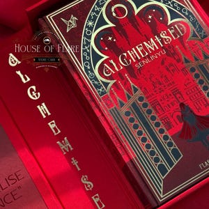 K&ouml;nnte beinhalten: Ein rotes Buch mit dem Titel "ALCHEMISED SENLINYU" in einer passenden Box. Das Buchcover zeigt ein detailliertes Design mit goldenen Akzenten und einer Silhouette einer Figur. Die Box und das Buch befinden sich auf einer roten Satin-Oberfl&auml;che. Der Text "HOUSE OF FLARE" ist ebenfalls sichtbar.