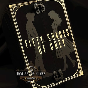 Może przedstawiać: Czarne pudełko ze złotymi zdobieniami i napisem "FIFTY SHADES OF GREY". Pudełko przedstawia sylwetki mężczyzny i kobiety. Pudełko ma zapięcie na zatrzask, widoczne są r&oacute;wnież słowa "HOUSE OF FLARE" i "ANDRE CARR".
