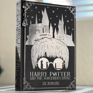 Puede incluir: Un libro negro de tapa dura con un dise&ntilde;o en plata de la escuela de magia de Hogwarts y Harry Potter con gafas. El t&iacute;tulo es "Harry Potter y la piedra filosofal" de J.K. Rowling.