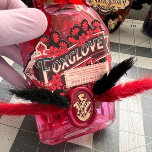 May include: A glass bottle with a red liquid and a label that reads "Foxglove". The bottle is decorated with red and black feathers and a red wax seal with a floral design. The label also includes the text "Ingredients: Foxglove Petals, Moonlit Dew Drops, Starlight Essence, Timeless Sandy & The Echoes of Destiny" and "2024 | 8.0 oz | 226".
