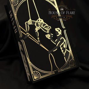 Może przedstawiać: Czarna książka ze złotymi detalami w stylu art deco i napisem "DESIRE" na grzbiecie. Okładka przedstawia złotą ilustrację dw&oacute;ch postaci. Książka nosi tytuł "HOUSE OF FLARE" i "ANDRE CARR".