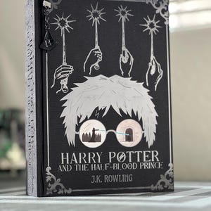 May include: A black hardcover book with a silver illustration of Harry Potter wearing glasses. The title "Harry Potter and the Half-Blood Prince" and the author "J.K. Rowling" are printed in silver on the cover.