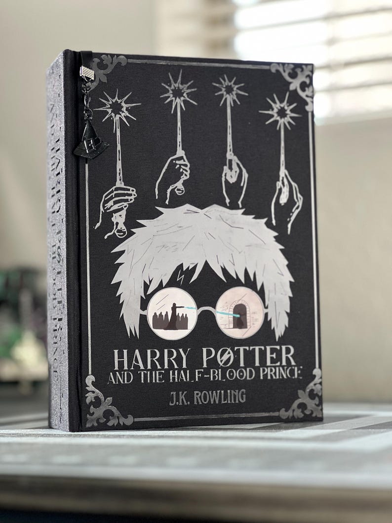 May include: A black hardcover book with a silver illustration of Harry Potter wearing glasses. The title "Harry Potter and the Half-Blood Prince" and the author "J.K. Rowling" are printed in silver on the cover.
