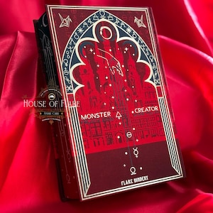 K&ouml;nnte beinhalten: Ein Hardcover-Buch mit rotem und schwarzem Einband, mit dem Text "MONSTER . CREATOR" und einer Illustration einer Stadt. Das Buch hat goldene Akzente und den Titel "FLARE BINDERY" unten. Das Buch liegt auf rotem Satinstoff.