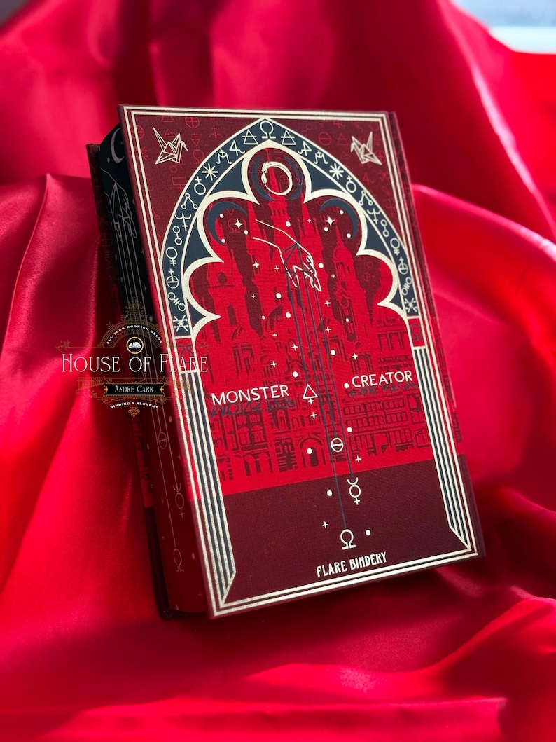 K&ouml;nnte beinhalten: Ein Hardcover-Buch mit rotem und schwarzem Einband, mit dem Text "MONSTER . CREATOR" und einer Illustration einer Stadt. Das Buch hat goldene Akzente und den Titel "FLARE BINDERY" unten. Das Buch liegt auf rotem Satinstoff.
