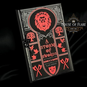 Può includere: Un libro nero intitolato "A Storm of Swords" di George R. R. Martin, con dettagli rossi e argento. La copertina presenta un emblema di leone, asce e disegni di alberi. Un ciondolo a forma di chiave è appeso al libro.