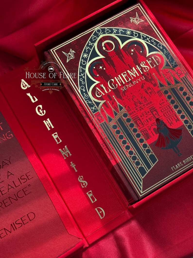 K&ouml;nnte beinhalten: Ein rotes Buch mit dem Titel "ALCHEMISED SENLINYU" in einer passenden Box. Das Buchcover zeigt ein detailliertes Design mit goldenen Akzenten und einer Silhouette einer Figur. Die Box und das Buch befinden sich auf einer roten Satin-Oberfl&auml;che. Der Text "HOUSE OF FLARE" ist ebenfalls sichtbar.
