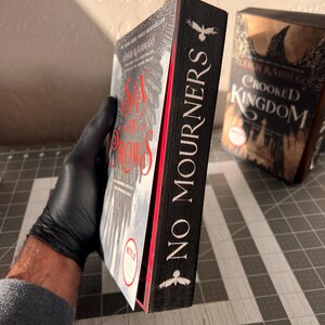 K&ouml;nnte beinhalten: Zwei Hardcover-B&uuml;cher mit schwarzen Einb&auml;nden und wei&szlig;em Text. Das Buch links tr&auml;gt den Titel "Six of Crows" und das Buch rechts den Titel "Crooked Kingdom". Beide B&uuml;cher haben einen schwarzen Raben auf dem R&uuml;cken mit dem Text "No Mourners" auf dem R&uuml;cken.