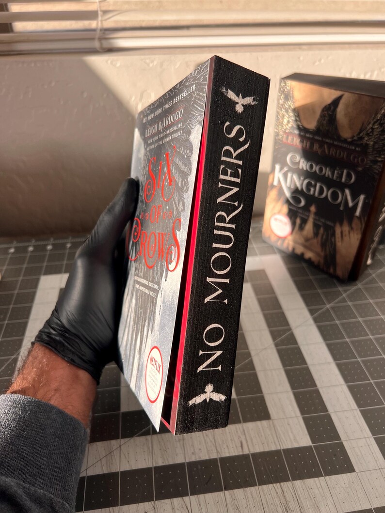 K&ouml;nnte beinhalten: Zwei Hardcover-B&uuml;cher mit schwarzen Einb&auml;nden und wei&szlig;em Text. Das Buch links tr&auml;gt den Titel "Six of Crows" und das Buch rechts den Titel "Crooked Kingdom". Beide B&uuml;cher haben einen schwarzen Raben auf dem R&uuml;cken mit dem Text "No Mourners" auf dem R&uuml;cken.