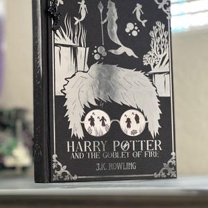 May include: A black book with a silver design featuring mermaids and a silver illustration of Harry Potter wearing glasses. The title of the book is "Harry Potter and the Goblet of Fire" by J.K. Rowling.