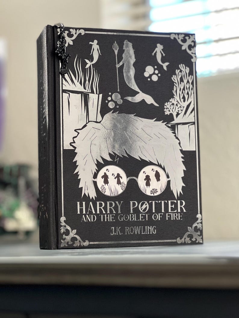 May include: A black book with a silver design featuring mermaids and a silver illustration of Harry Potter wearing glasses. The title of the book is "Harry Potter and the Goblet of Fire" by J.K. Rowling.