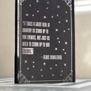 Puede incluir: Un cuaderno negro con una cita en plata grabada de Albus Dumbledore: "Se necesita mucha valent&iacute;a para enfrentarse a nuestros enemigos, pero igual para enfrentarse a nuestros amigos." El cuaderno tiene un borde plateado y un patr&oacute;n de estrellas plateadas.