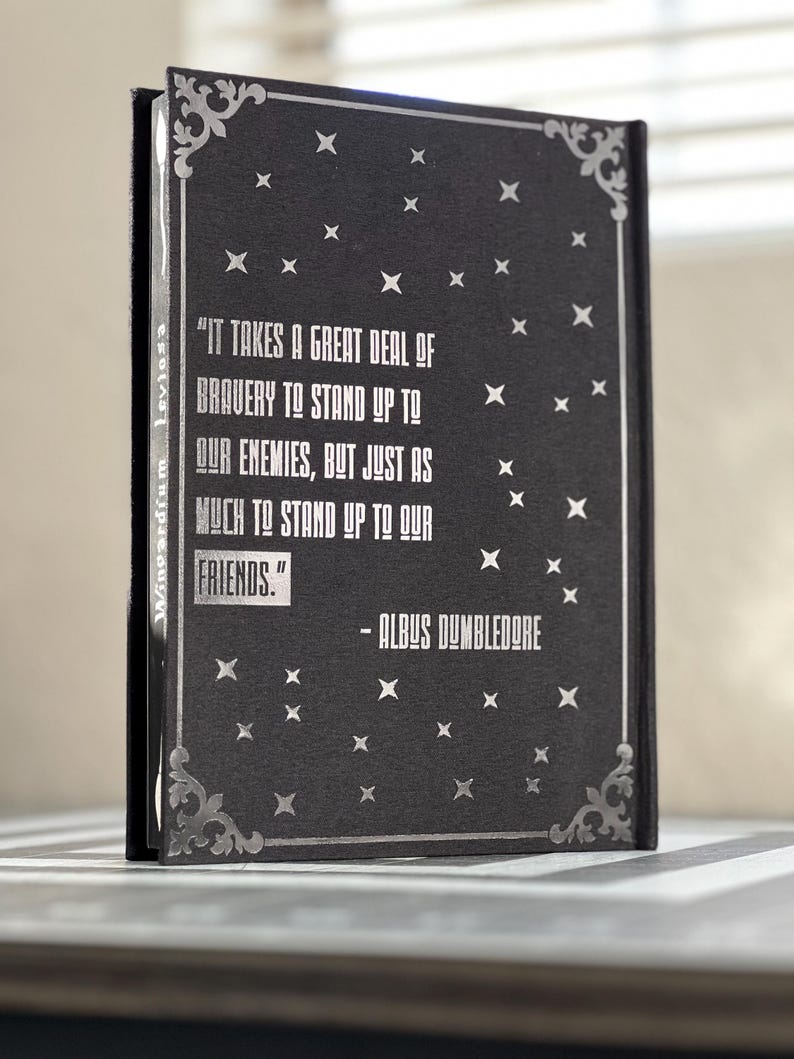 Puede incluir: Un cuaderno negro con una cita en plata grabada de Albus Dumbledore: "Se necesita mucha valent&iacute;a para enfrentarse a nuestros enemigos, pero igual para enfrentarse a nuestros amigos." El cuaderno tiene un borde plateado y un patr&oacute;n de estrellas plateadas.