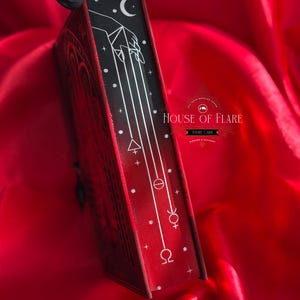 K&ouml;nnte beinhalten: Ein rotes Buch mit schwarzem Buchr&uuml;cken, das einen wei&szlig;en Mond, eine Handillustration und Himmelszeichen zeigt. Der Einband des Buches weist aufwendige Pr&auml;gemuster auf. Der Text "HOUSE OF FLARE" ist auf dem Einband zu sehen.