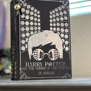 May include: A black hardcover book with a silver design of Harry Potter wearing glasses. The title of the book is "Harry Potter and the Order of the Phoenix" by J.K. Rowling.
