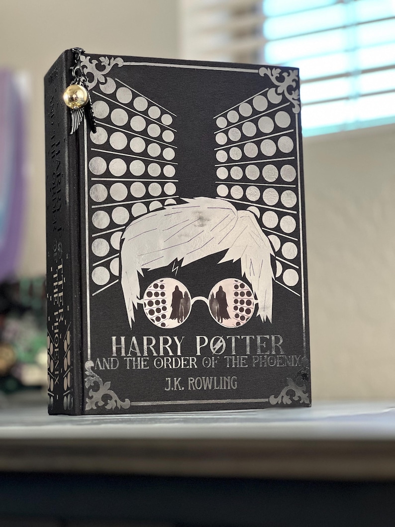May include: A black hardcover book with a silver design of Harry Potter wearing glasses. The title of the book is "Harry Potter and the Order of the Phoenix" by J.K. Rowling.