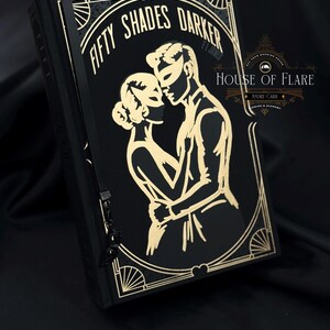 Może przedstawiać: Czarna książka ze złotymi literami i złotym wzorem w stylu art deco. Okładka książki głosi "FIFTY SHADES DARKER" i zawiera ilustrację obejmującej się pary, oboje w maskach. Książka ma czarną wstążkę jako zakładkę.