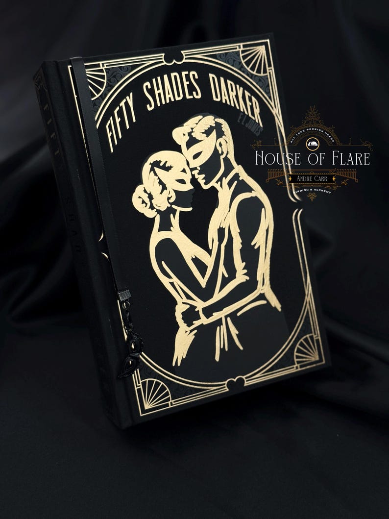 Może przedstawiać: Czarna książka ze złotymi literami i złotym wzorem w stylu art deco. Okładka książki głosi "FIFTY SHADES DARKER" i zawiera ilustrację obejmującej się pary, oboje w maskach. Książka ma czarną wstążkę jako zakładkę.