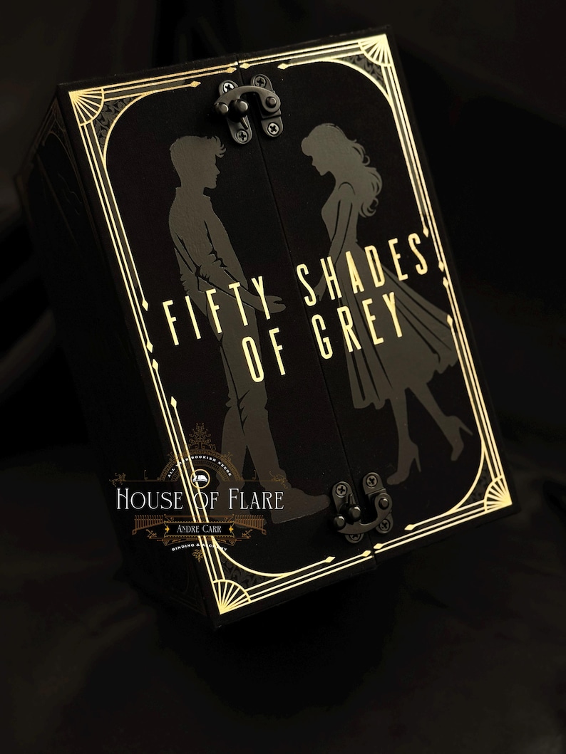 Może przedstawiać: Czarne pudełko ze złotymi zdobieniami i napisem "FIFTY SHADES OF GREY". Pudełko przedstawia sylwetki mężczyzny i kobiety. Pudełko ma zapięcie na zatrzask, widoczne są r&oacute;wnież słowa "HOUSE OF FLARE" i "ANDRE CARR".