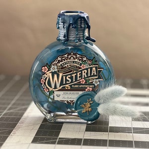 May include: A blue glass bottle with a wax seal and a label that reads "Wisteria Flare Apothecary" and "Extend Life Through Death". The bottle is filled with a blue liquid and has a white feather on the side.