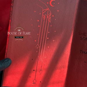 K&ouml;nnte beinhalten: Offenes Buch mit roten Seiten und einer detaillierten Illustration. Die linke Seite zeigt eine Hand mit Linien, einen Halbmond und Symbole. Das Buch tr&auml;gt die Aufschrift "HOUSE OF FLARE" und "ANDRE CARR".