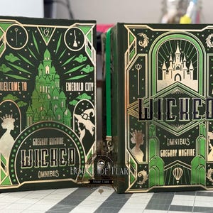 以下が含まれることがあります： 緑と金の箔のデザインが施された2冊のハードカバー本。左の本には「Welcome to Emerald City」と書かれており、緑色の城が描かれています。右の本には「Wicked Omnibus」と書かれており、城のイラストが描かれています。どちらの本にも著者の名前が書かれています。