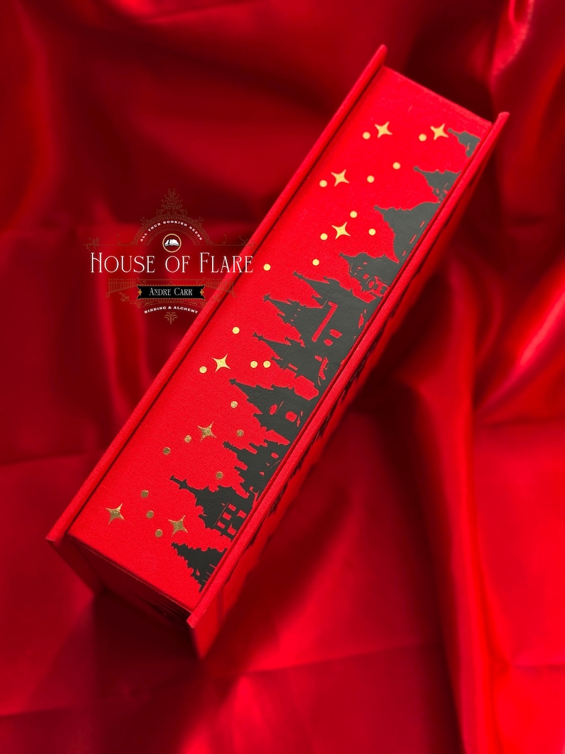 K&ouml;nnte beinhalten: Eine rote Schachtel mit einem schwarzen Silhouette-Design einer Stadtlandschaft und goldenen Sternen. Auf der Schachtel steht "HOUSE OF FLARE" und "ANDRE CARR". Die Schachtel liegt auf einer roten Satin-Oberfl&auml;che.