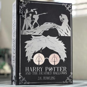 May include: A black hardcover book with a silver illustration of Harry Potter and the Deathly Hallows on the cover. The title "Harry Potter and the Deathly Hallows" and the author "J.K. Rowling" are printed in silver on the cover.