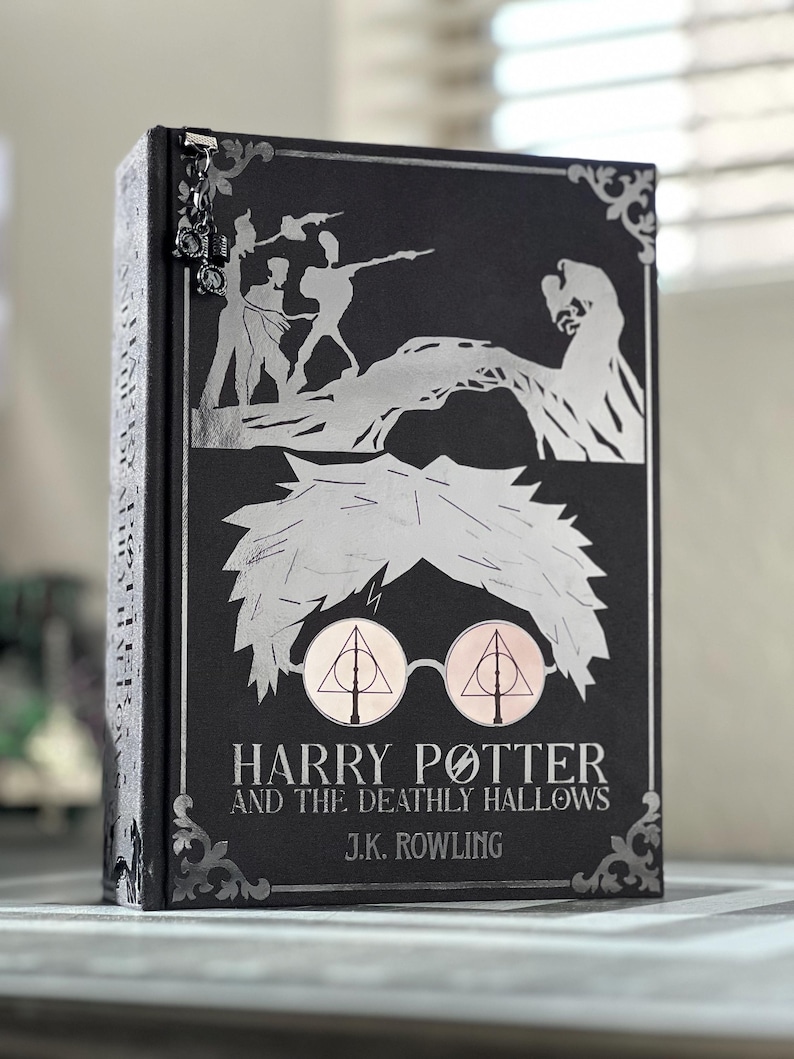 May include: A black hardcover book with a silver illustration of Harry Potter and the Deathly Hallows on the cover. The title "Harry Potter and the Deathly Hallows" and the author "J.K. Rowling" are printed in silver on the cover.