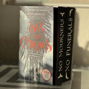 K&ouml;nnte beinhalten: Zwei Taschenb&uuml;cher, die &uuml;bereinander gestapelt sind. Das obere Buch tr&auml;gt den Titel "Six of Crows" von Leigh Bardugo. Das untere Buch tr&auml;gt den Titel "No Mourners, No Funerals". Beide B&uuml;cher haben einen schwarzen Hintergrund mit wei&szlig;em Text.