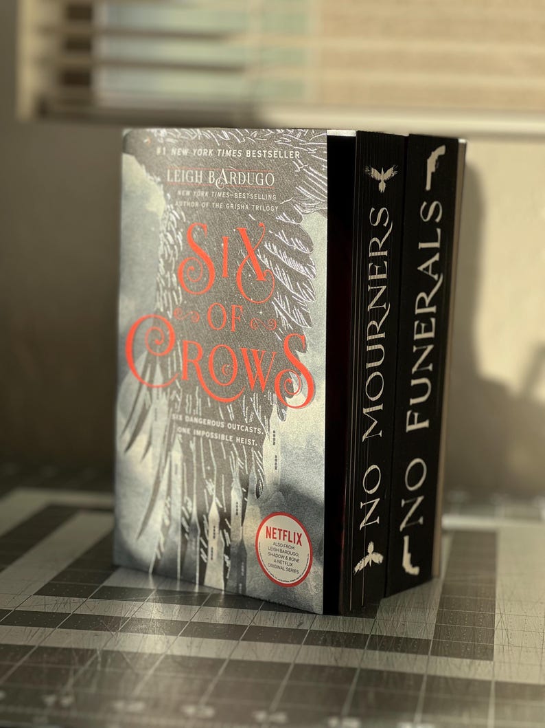 K&ouml;nnte beinhalten: Zwei Taschenb&uuml;cher, die &uuml;bereinander gestapelt sind. Das obere Buch tr&auml;gt den Titel "Six of Crows" von Leigh Bardugo. Das untere Buch tr&auml;gt den Titel "No Mourners, No Funerals". Beide B&uuml;cher haben einen schwarzen Hintergrund mit wei&szlig;em Text.