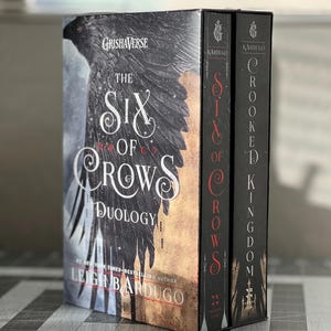 K&ouml;nnte beinhalten: Ein zweib&auml;ndiges Box-Set der Six of Crows-Duologie von Leigh Bardugo. Die B&uuml;cher tragen die Titel "Six of Crows" und "Crooked Kingdom". Die Cover zeigen einen schwarzen Raben vor einer Stadtkulisse.