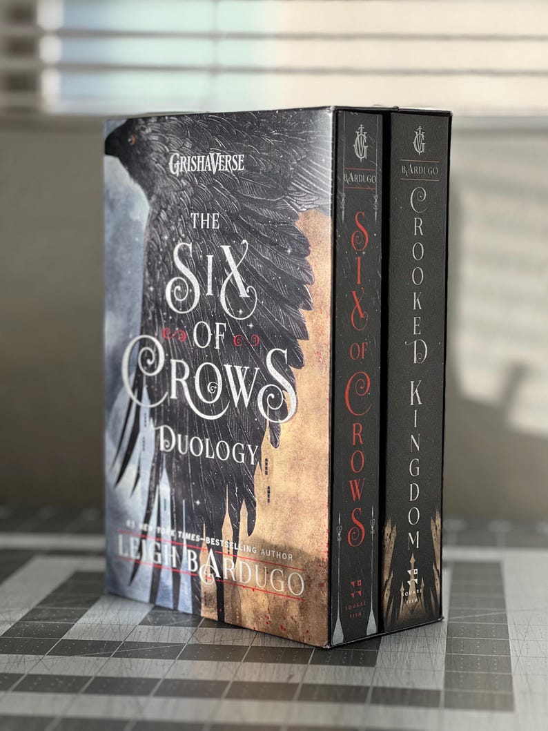 K&ouml;nnte beinhalten: Ein zweib&auml;ndiges Box-Set der Six of Crows-Duologie von Leigh Bardugo. Die B&uuml;cher tragen die Titel "Six of Crows" und "Crooked Kingdom". Die Cover zeigen einen schwarzen Raben vor einer Stadtkulisse.