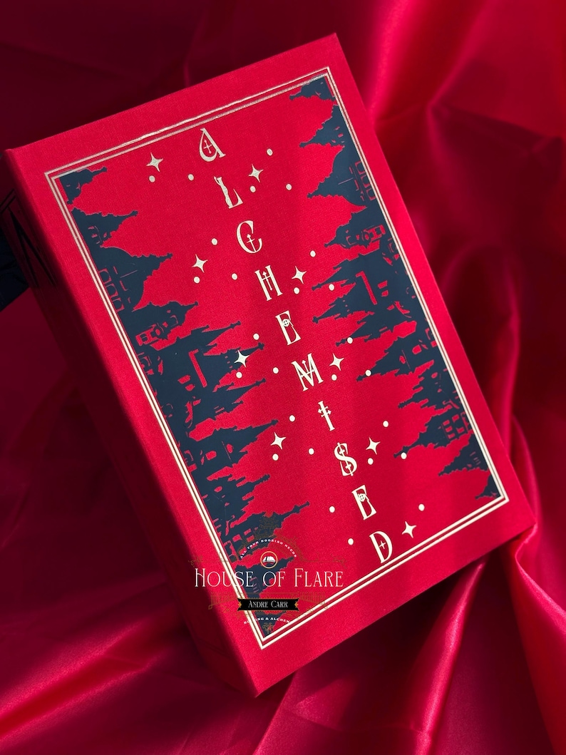 K&ouml;nnte beinhalten: Ein rotes Buch mit goldenem Rand und dem Titel "ALCHEMISED" in Wei&szlig;. Das Cover zeigt ein dunkelblaues Silhouette-Design von Geb&auml;uden und Sternen. Das Buch liegt auf einem roten Satinhintergrund, mit dem Logo "HOUSE OF FLARE" sichtbar.