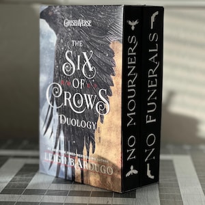 K&ouml;nnte beinhalten: Ein zweib&auml;ndiges Box-Set der Grishaverse-Reihe von Leigh Bardugo, die "Six of Crows"-Duologie. Die B&uuml;cher sind schwarz mit silberner Beschriftung und zeigen einen Raben auf dem vorderen Umschlag.