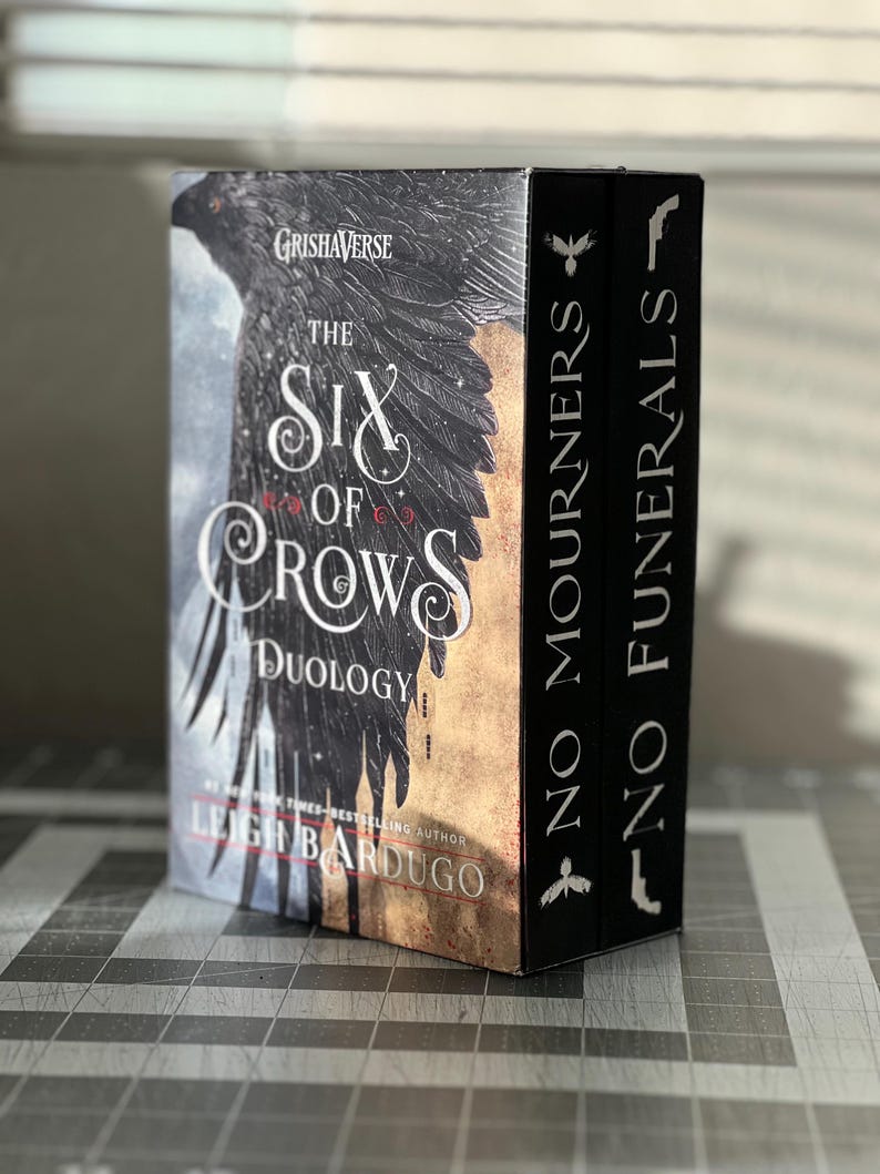K&ouml;nnte beinhalten: Ein zweib&auml;ndiges Box-Set der Grishaverse-Reihe von Leigh Bardugo, die "Six of Crows"-Duologie. Die B&uuml;cher sind schwarz mit silberner Beschriftung und zeigen einen Raben auf dem vorderen Umschlag.
