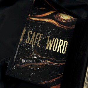 Może przedstawiać: Czarna książka z marmurowym wzorem i złotymi literami z napisem "SAFE WORD". Książka znajduje się w czarnym pudełku z napisem "HOUSE OF FLARE" i "ANDRE CARR" na okładce. Książka ma czarną wstążkę.