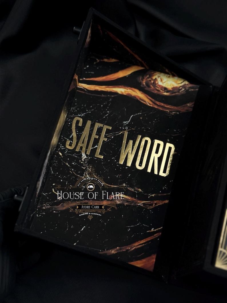 Może przedstawiać: Czarna książka z marmurowym wzorem i złotymi literami z napisem "SAFE WORD". Książka znajduje się w czarnym pudełku z napisem "HOUSE OF FLARE" i "ANDRE CARR" na okładce. Książka ma czarną wstążkę.