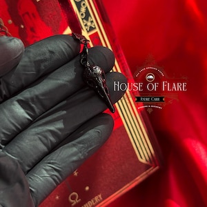 K&ouml;nnte beinhalten: Schwarzer Rabensch&auml;del-Anh&auml;nger mit filigranen Details, vor rotem Hintergrund. Der Anh&auml;nger h&auml;ngt an einer feinen Kette. Ein rotes Buch mit goldenen Akzenten und dem Text "HOUSE OF FLARE" ist ebenfalls zu sehen.