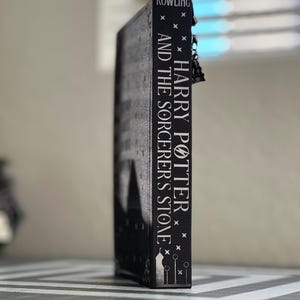 Puede incluir: Un libro negro con el t&iacute;tulo "Harry Potter y la piedra filosofal" de J.K. Rowling en el lomo.