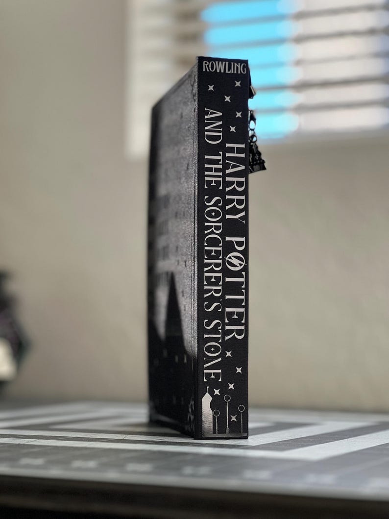 Puede incluir: Un libro negro con el t&iacute;tulo "Harry Potter y la piedra filosofal" de J.K. Rowling en el lomo.