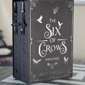 May include: A black hardcover book with silver lettering. The title is "The Six of Crows Duology" by Leigh Bardugo. The cover features a decorative design with silver crows.