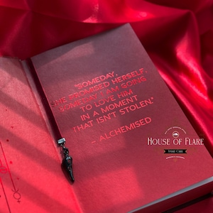 K&ouml;nnte beinhalten: Offenes rotes Buch mit schwarzem Lesezeichen und Anh&auml;nger. Die Buchseiten zeigen roten Text: "SOMEDAY, SHE PROMISED HERSELF, SOMEDAY I AM GOING TO LOVE HIM IN A MOMENT THAT ISN'T STOLEN." Das Buch tr&auml;gt den Titel "ALCHEMISED".