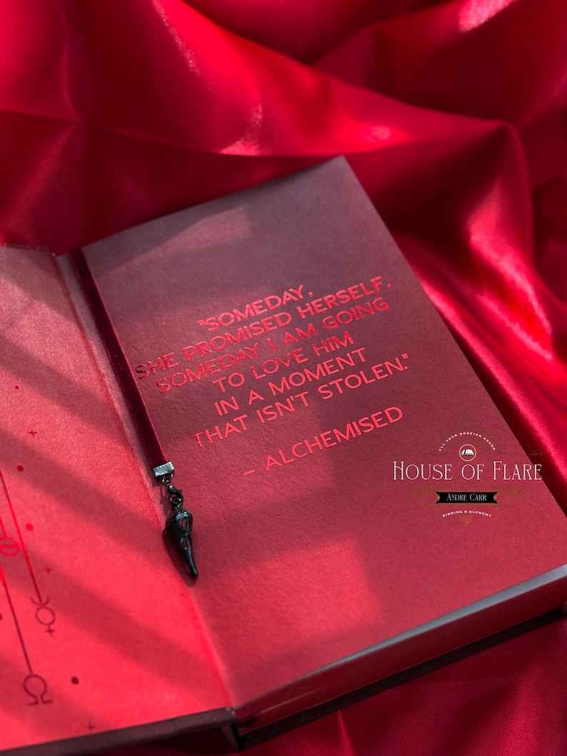 K&ouml;nnte beinhalten: Offenes rotes Buch mit schwarzem Lesezeichen und Anh&auml;nger. Die Buchseiten zeigen roten Text: "SOMEDAY, SHE PROMISED HERSELF, SOMEDAY I AM GOING TO LOVE HIM IN A MOMENT THAT ISN'T STOLEN." Das Buch tr&auml;gt den Titel "ALCHEMISED".