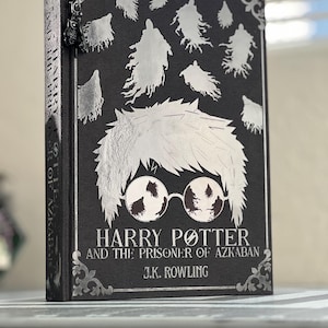 May include: A black hardcover book with silver foil lettering. The title is "Harry Potter and the Prisoner of Azkaban" by J.K. Rowling. The cover features a silver silhouette of Harry Potter with glasses and Dementors flying around him.