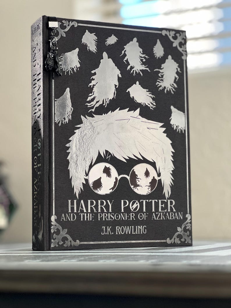May include: A black hardcover book with silver foil lettering. The title is "Harry Potter and the Prisoner of Azkaban" by J.K. Rowling. The cover features a silver silhouette of Harry Potter with glasses and Dementors flying around him.